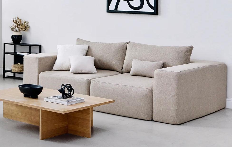 A modern living room with a beige couch adorned with white and beige throw pillows. In front of the couch is a wooden coffee table displaying books and a decorative black sculpture. A black shelf stands in the corner, holding a plant and baskets. White wall art hangs above.