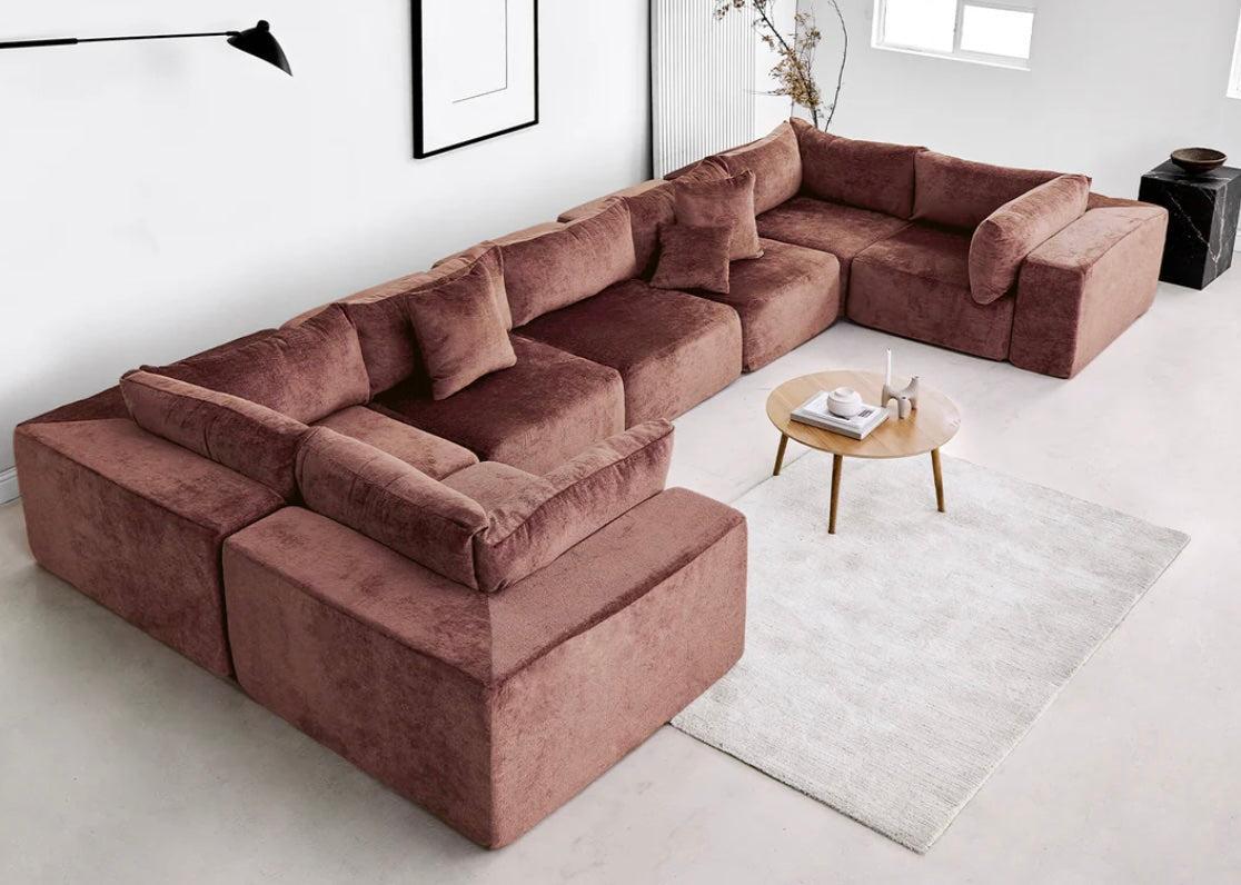 A spacious living room featuring a large, plush, brown sectional sofa with multiple cushions. The seating area is complemented by a circular wooden coffee table displaying a white vase and small decorations, placed on a light-colored rug. A minimalist wall lamp and framed art are on the walls.