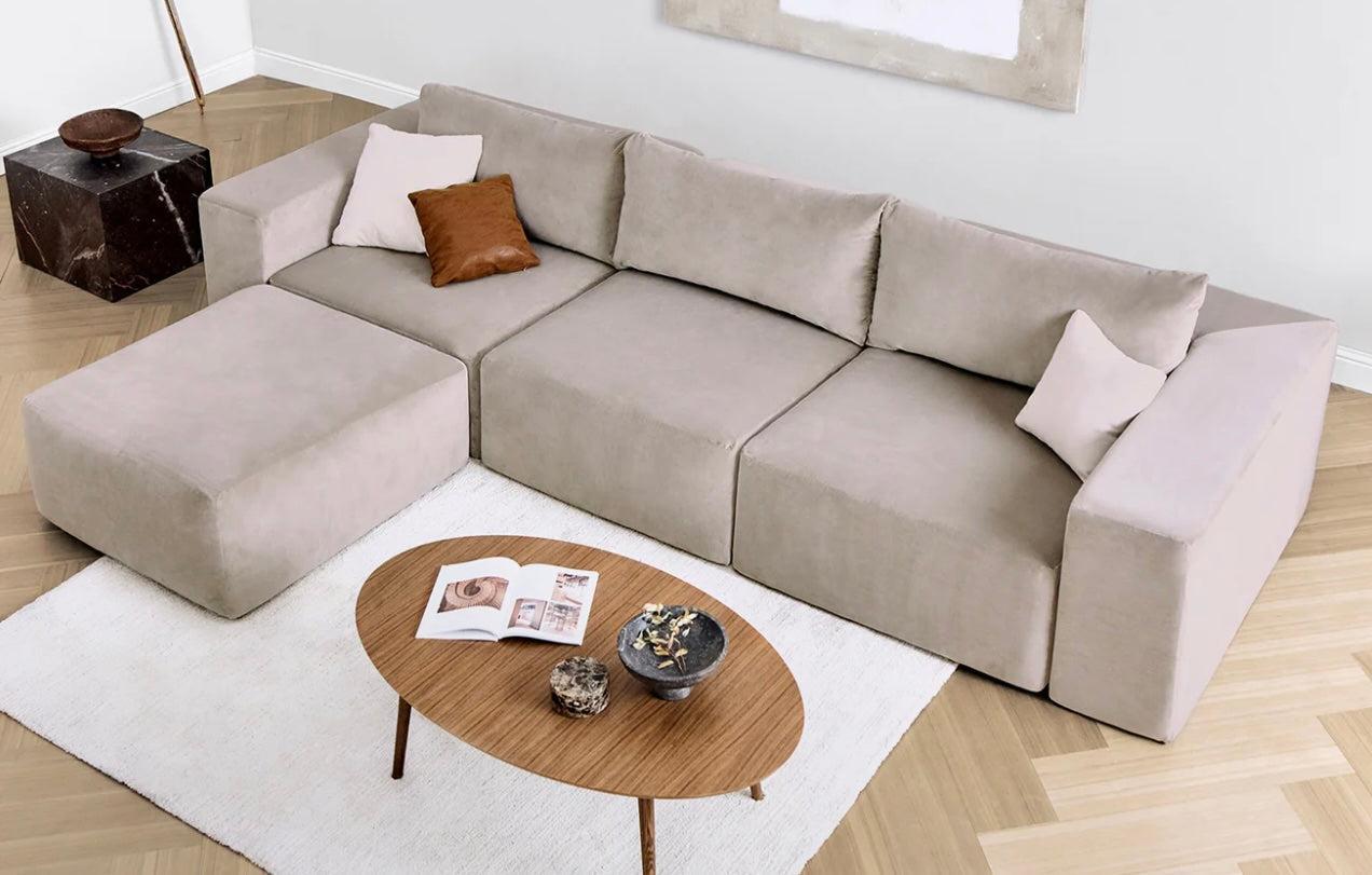 A modern living room setup featuring a light beige sectional sofa with matching cushions, a brown leather pillow, and a white one. A wooden coffee table holds an open book and decorative items atop a white rug on light wood flooring. A marble accent table is in the corner.