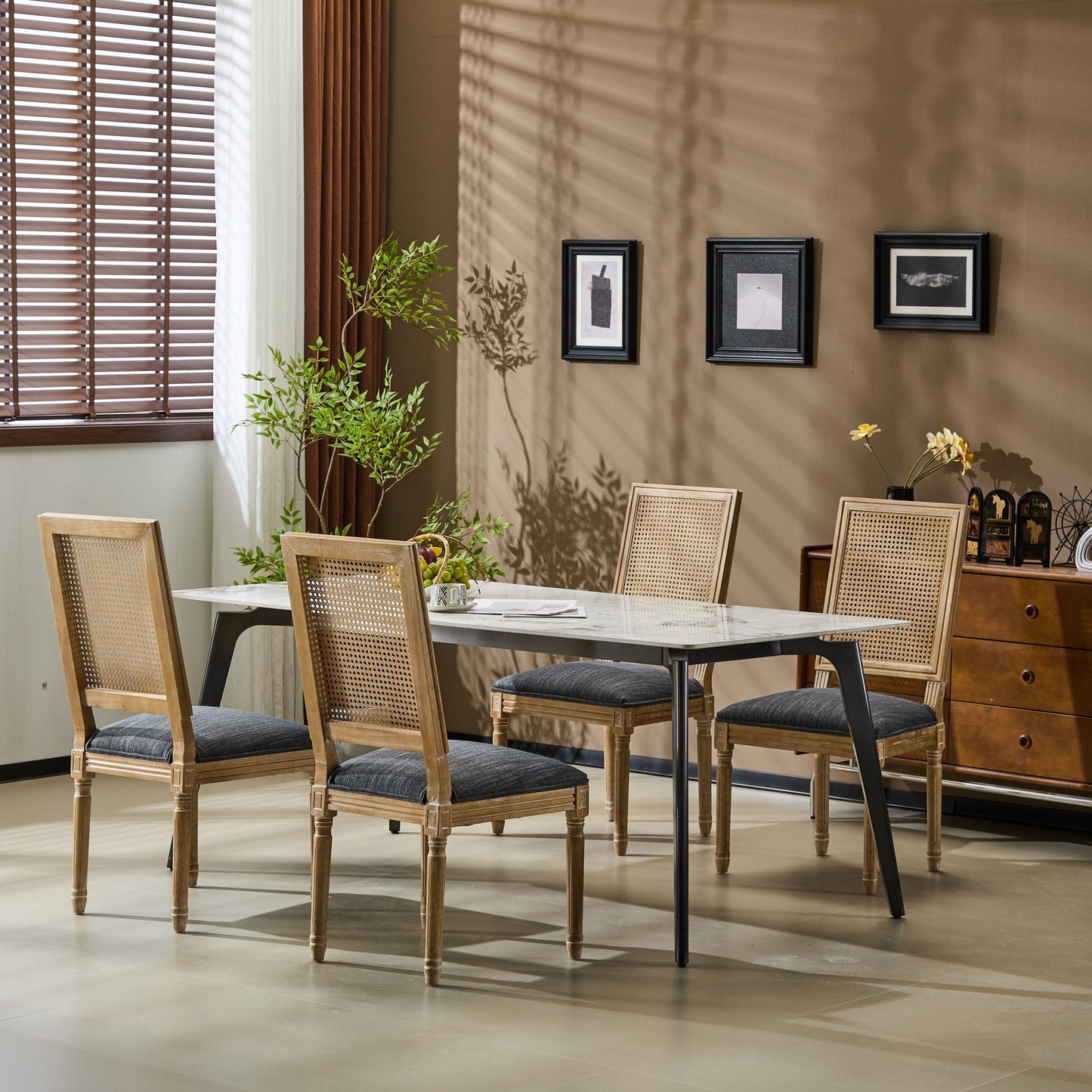 A modern dining room with a rectangular table, four French Country Dining Chairs with rectangular cane backrests and smooth upholstered seating, a potted plant, and framed art on a beige wall. Sunlight streams through window blinds, casting shadows.