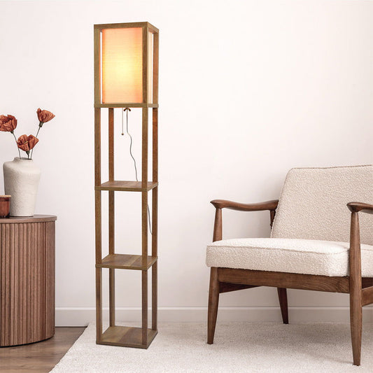 The Pinnacle Shelf Floor Lamp For Bedroom / Living Room, Long Shade - Natural Wood / White stands beside a light beige armchair and wooden side table with vases in a minimalist living room, casting a cozy warm glow.