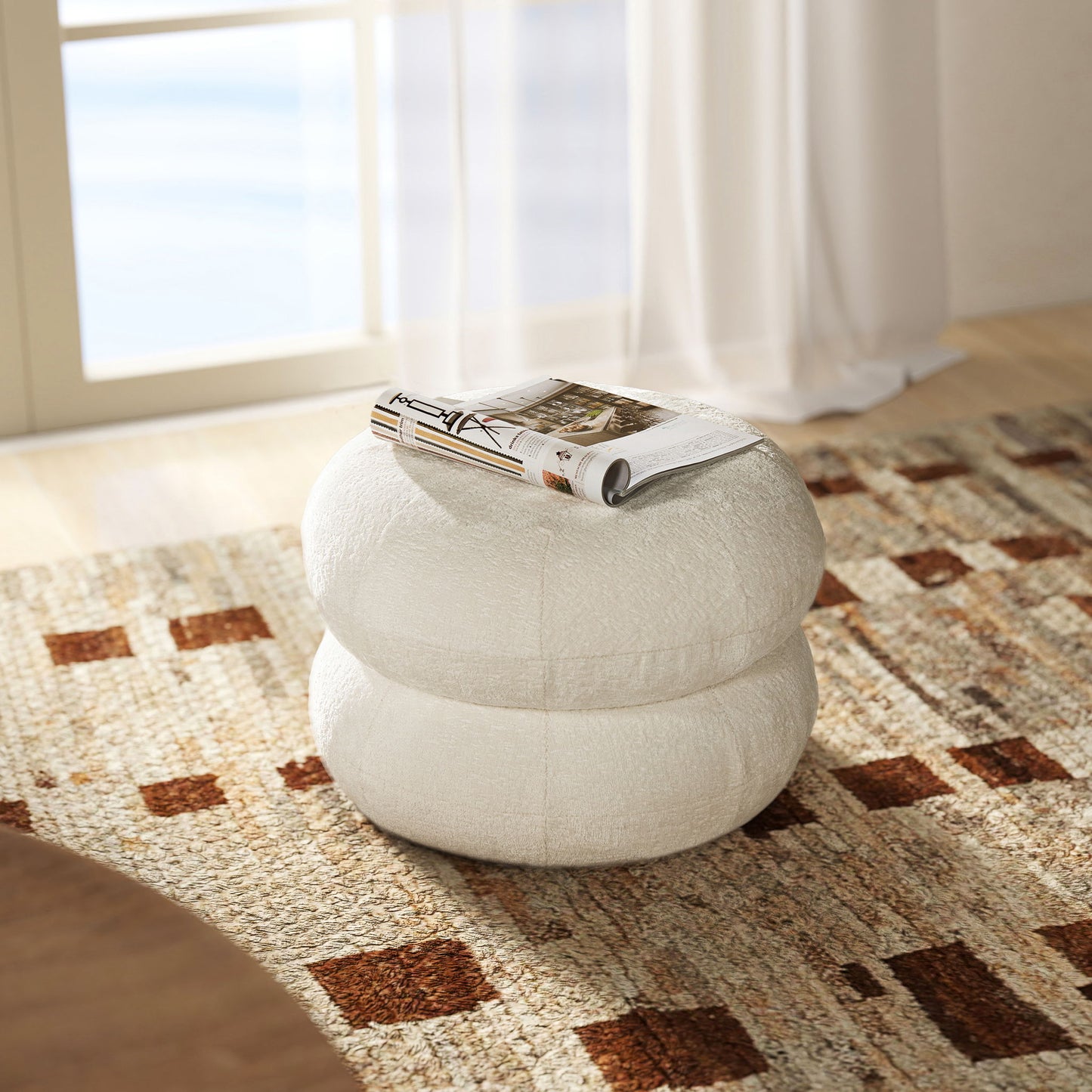 The Nest - Low Round Upholstered Ottoman, featuring a white textured surface, sits on an earth-toned patterned rug by the window. A rolled magazine rests on top as sunlight filters through sheer curtains in the background.