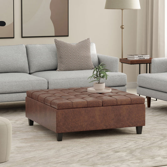 A modern living room with a light gray sofa, the Harrison Upholstered Coffee Table Storage Ottoman in brown, a potted plant atop it, abstract wall art, a side table with books, and a floor lamp—all in soothing neutral tones.