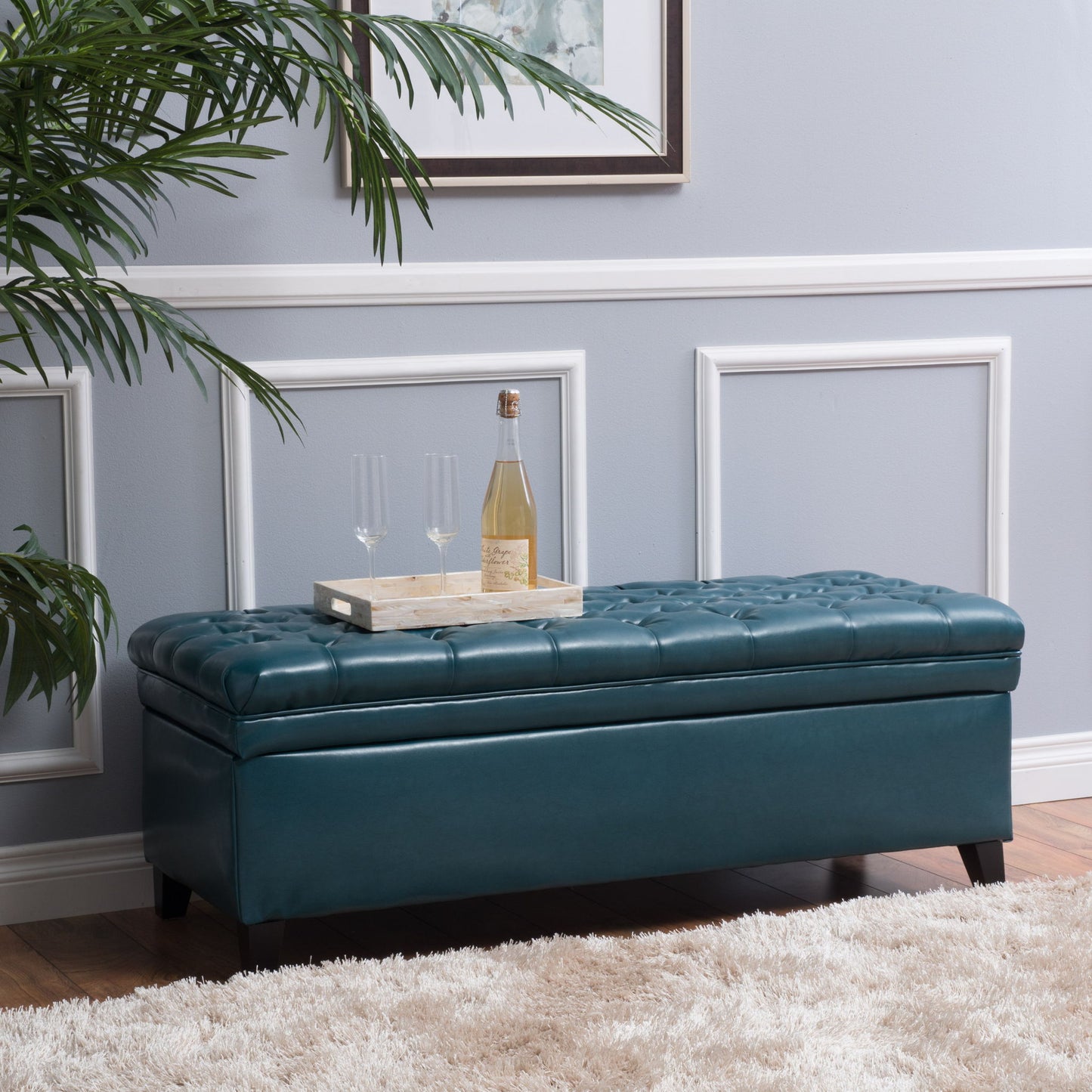The Juliana Storage Ottoman with Birch Legs in teal sits against a light gray wall with framed molding. A wooden tray with a wine bottle and two glasses rests on top, alongside a green plant and a white rug.
