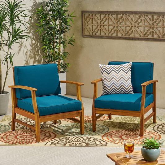 Two Perla Contemporary Club Chairs with water-resistant teal cushions sit on a patterned rug; one features a zigzag pillow. A wooden table with a drink is in front, surrounded by green potted plants and wall art in the background.