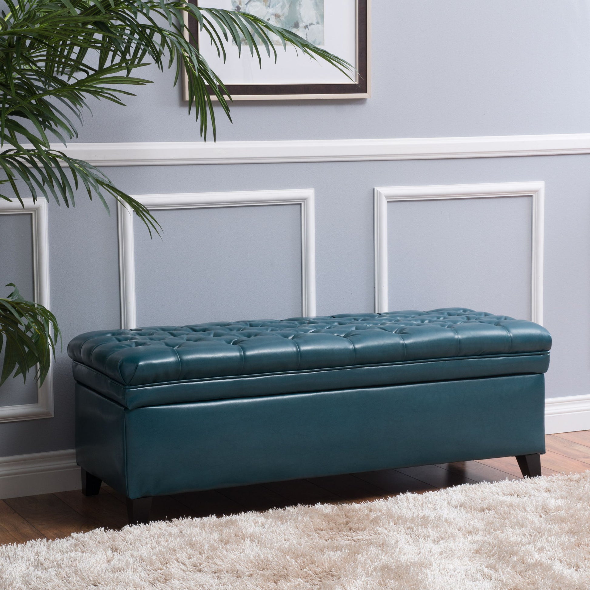 The Juliana Storage Ottoman With Birch Legs in teal leather sits against a light gray wall with white molding, next to a potted plant and a cream-colored rug.