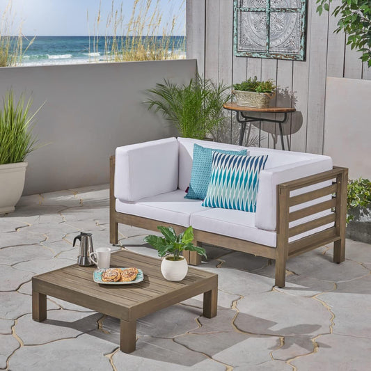 Create a serene patio with the Oana 3 Piece Love Seat Set With Coffee Table, featuring a wooden loveseat with white cushions, blue patterned pillows, and a low table perfect for enjoying pastries and coffee by the ocean.