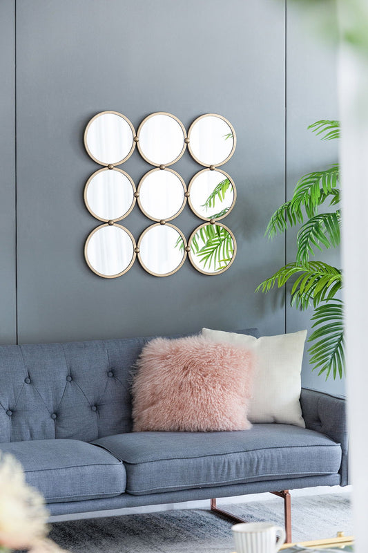 A modern living room features a blue tufted sofa, pink and white pillows, and a grid of Contemporary Decorative Mirrors in a minimalist champagne finish on a gray wall reflecting a large green plant.