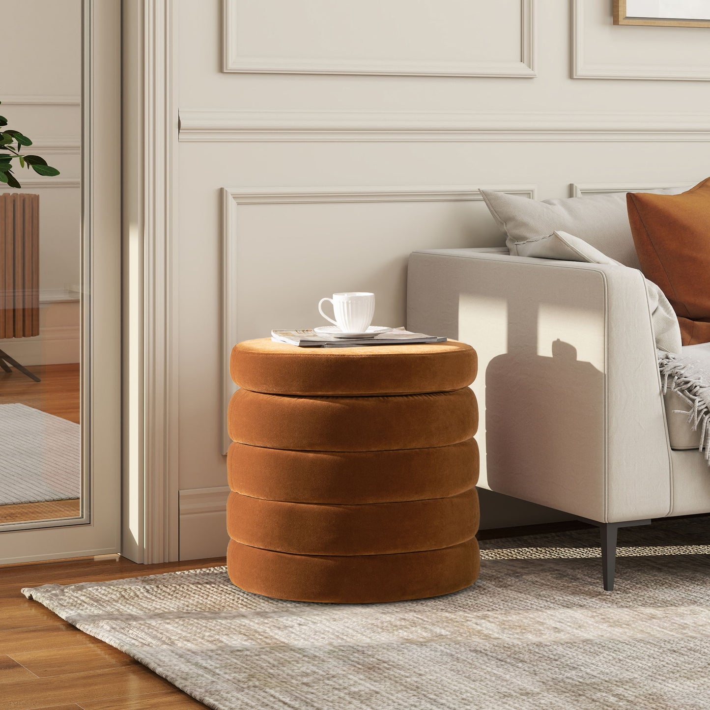 The Fuji Oversized Round Storage Ottoman in warm brown velvet serves as a side table, holding a white cup and magazine next to a beige sofa in a bright, modern living room with neutral decor.