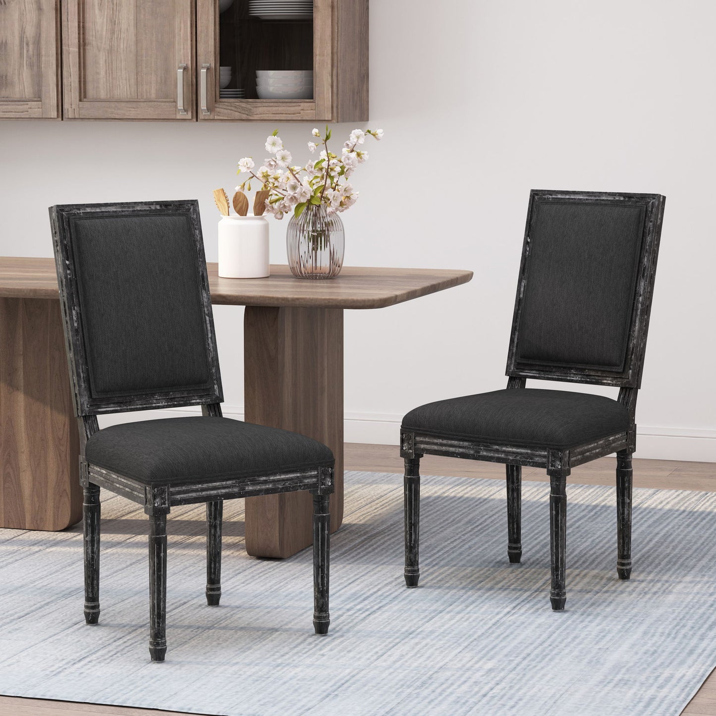Two French Country Style Dining Chairs with upholstered seats and fluted legs, featuring elegant black fabric and distressed wood frames, sit side by side on a light striped rug in a modern dining room with a wooden table and cabinets.