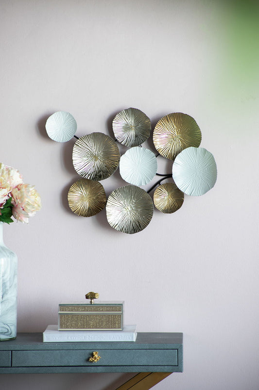 The Contemporary Metal Wall Decor Accent in golden and white features clustered circular discs with textured lines, shown mounted on a light pink wall above a table with flowers and stacked books.