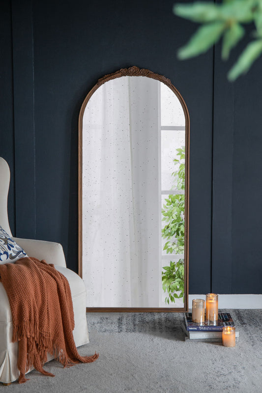 A Hand Carved Rose Antique Mirror with a large brown arch frame leans against a dark blue wall, reflecting greenery outside. Nearby, a cream armchair with an orange throw and lit candles on stacked books create a cozy scene.