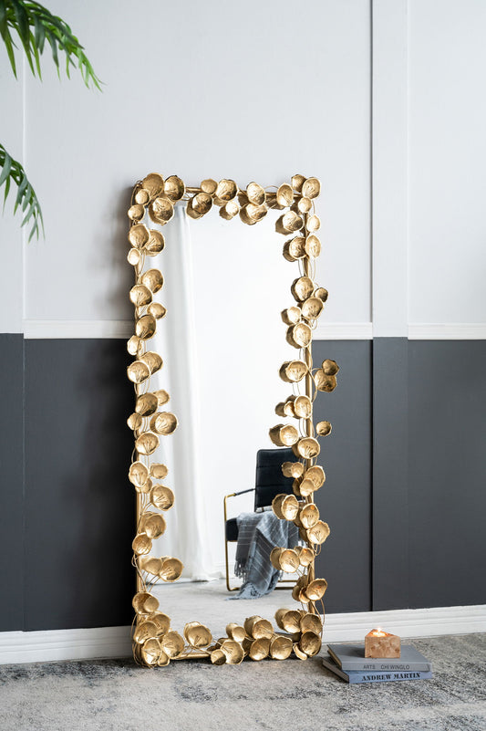 The Full Length Mirror with en Leaf Accents in gold, designed for living rooms or bedrooms, leans against a two-tone wall in a modern room with gray carpet and a plant partly visible on the left.