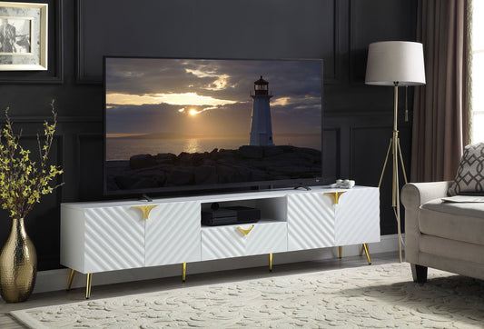 A modern living room features the Gaines High Gloss TV Stand in white, a flat-screen TV with a lighthouse sunset scene, a floor lamp, beige sofa, patterned rug, and decorative plant set against dark paneled walls.