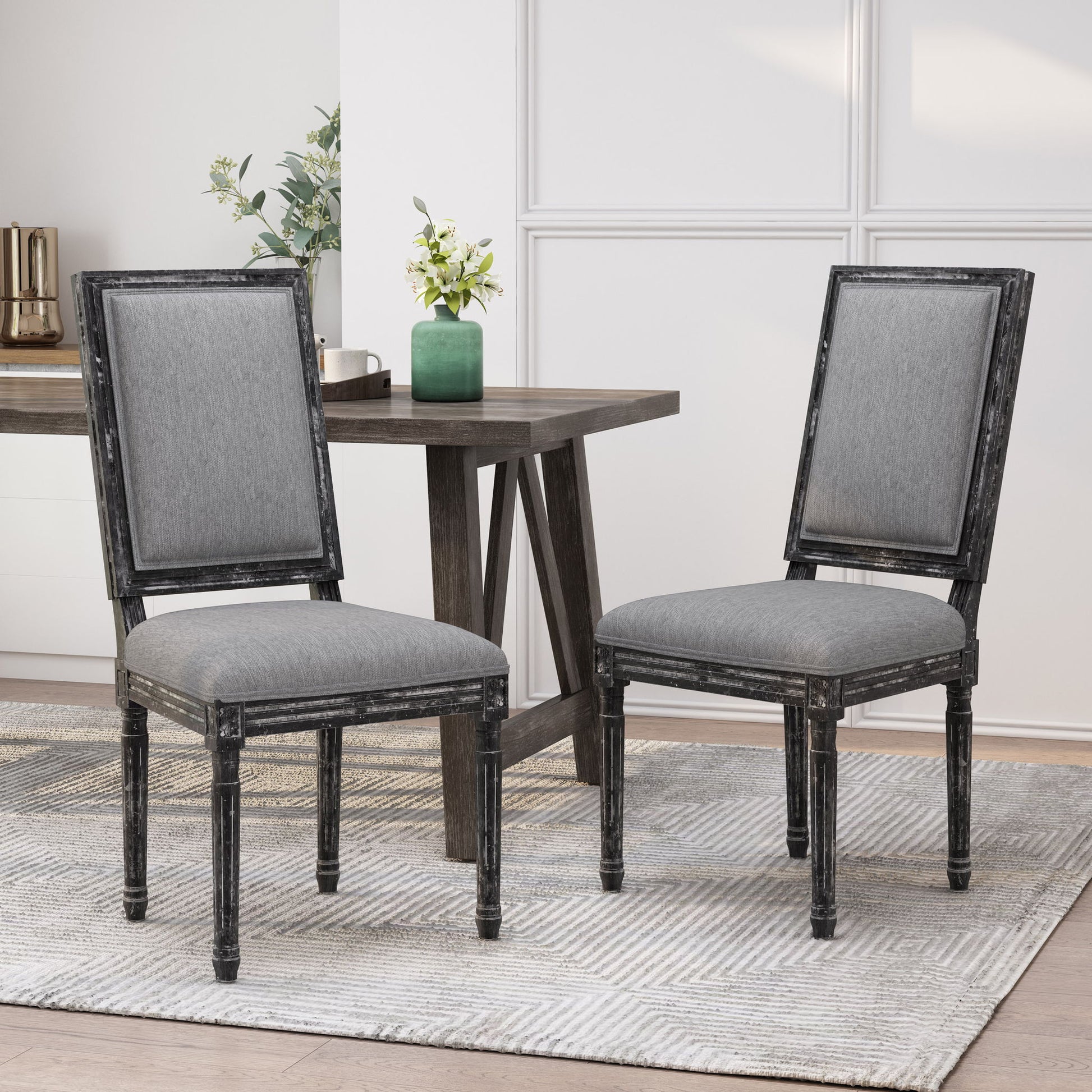 Two French Country Style Dining Chairs with upholstered seating and fluted legs are set on a patterned rug beside a dark wood table topped with a green vase of white flowers in a bright, modern dining room.