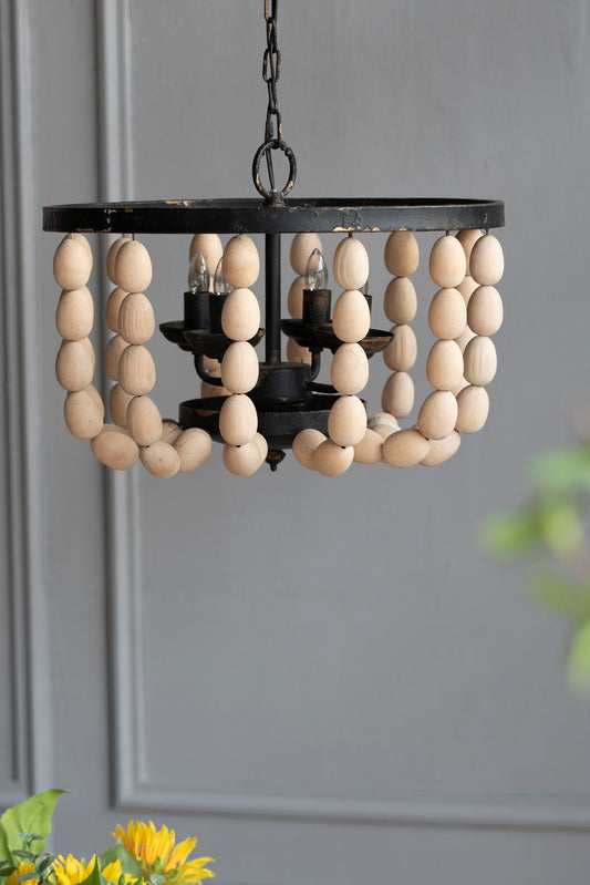 The 4 Light Wood Chandelier, a hanging light fixture with an adjustable chain in cream (bulb not included), is showcased against a gray paneled wall, with green leaves and yellow flowers in the bottom left corner.