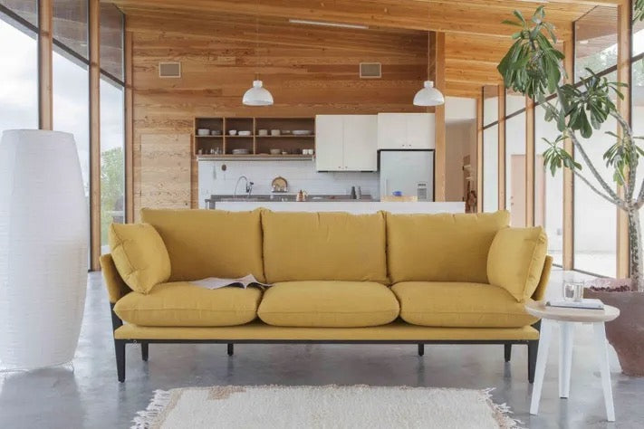 A modern living room with a mustard yellow sofa in the center, a white floor lamp on the left, and a small white side table with a plant on the right. The background features an open kitchen with wooden cabinets and a large potted plant.