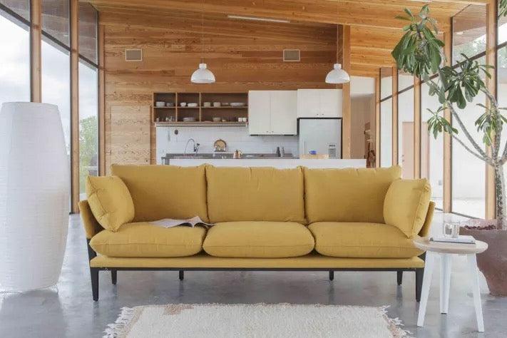 A modern living room with a mustard yellow sofa in the center, a white floor lamp on the left, and a small white side table with a plant on the right. The background features an open kitchen with wooden cabinets and a large potted plant.