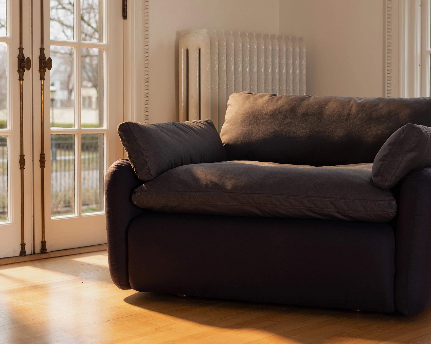 A cozy, dark gray armchair bathed in sunlight sits next to a set of French doors. The warm wooden floor complements the inviting atmosphere of the room. Outside, trees and a fence can be seen through the glass panes of the doors.