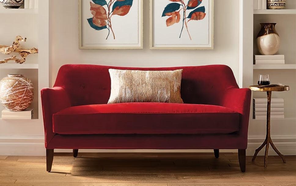 A stylish living room features a red velvet loveseat with a beige throw pillow in the center. Above the loveseat, two framed botanical prints with red and blue leaves are hung on a white wall. A small side table to the right holds a glass of red wine.