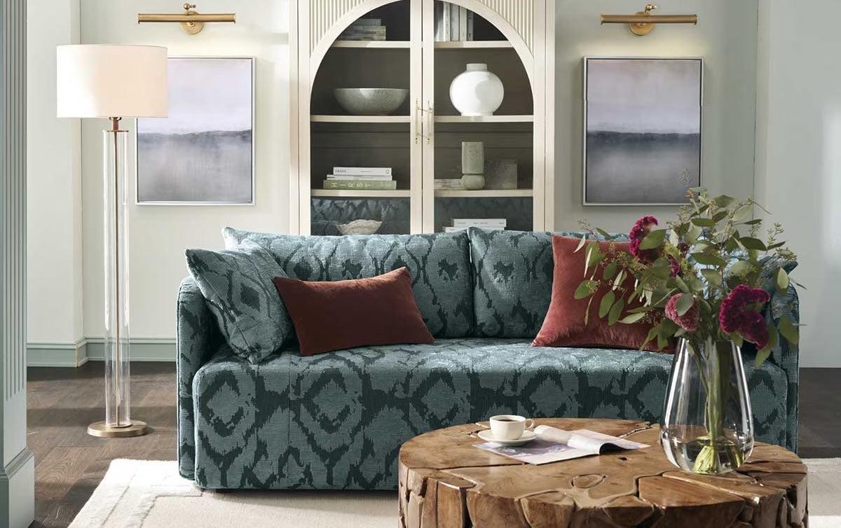 A stylish living room features a blue patterned sofa with red and blue throw pillows, a wooden coffee table with a vase of flowers, a floor lamp, and a tall bookshelf in the background. Two abstract paintings flank the bookshelf.