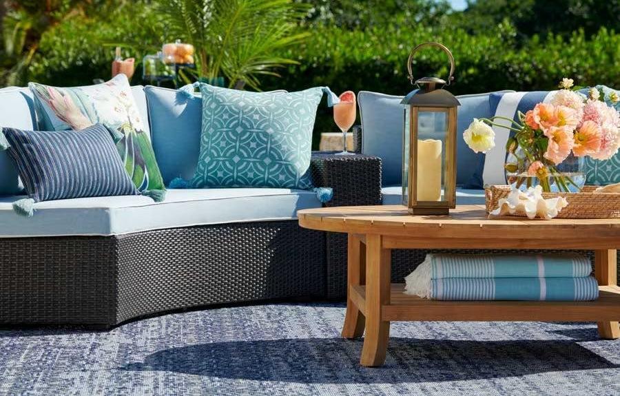 A cozy outdoor seating area with a wicker sectional sofa adorned with blue and white patterned cushions. A wooden coffee table is set with a lantern, a bouquet of flowers, and rolled-up towels. The scene is bright and inviting, set against a lush green background.