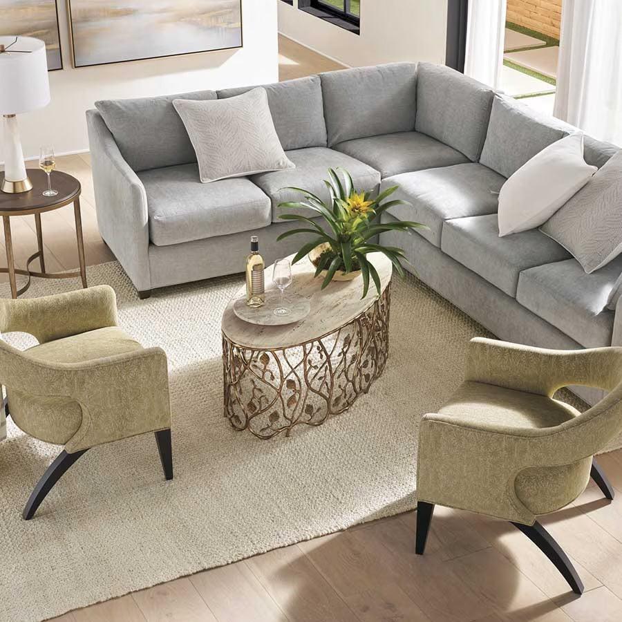 A cozy living room with a light gray sectional sofa and two olive green armchairs arranged around a glass-top coffee table with a decorative metal base. The table features a potted plant and a bottle of wine with a glass. Beige rug covers the wooden floor.
