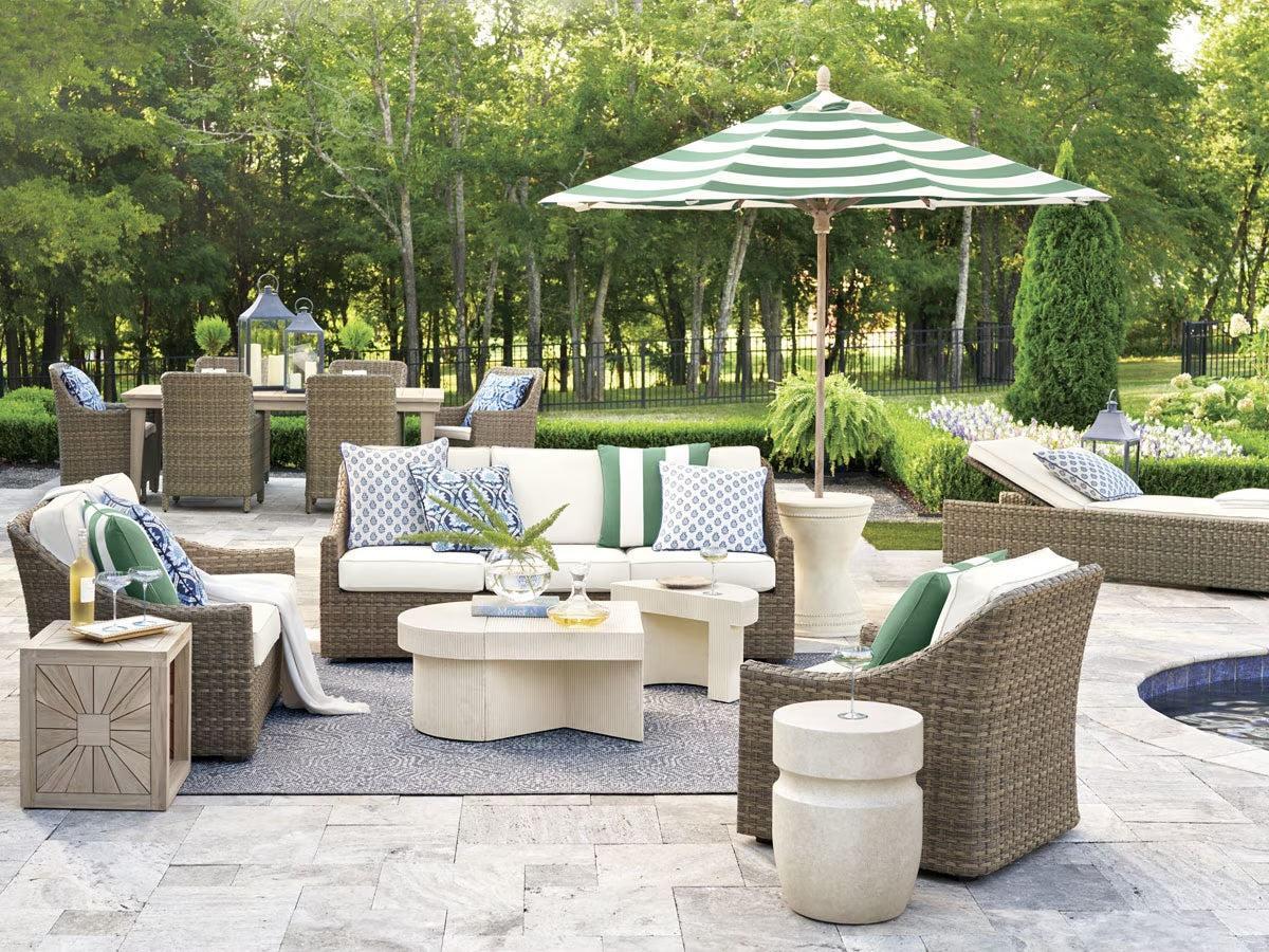 A stylish outdoor patio features wicker furniture with green and white cushions, a low white coffee table, decorative pillows, and a green striped umbrella. Surrounded by lush greenery, the setting includes a pool and tall trees in the background.