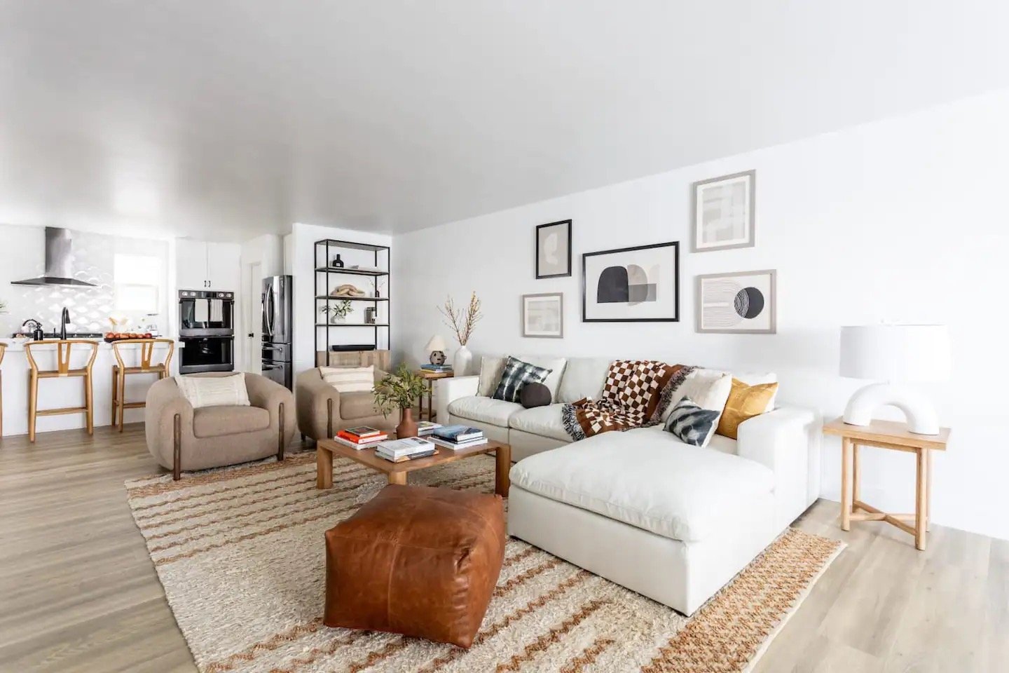 A modern living room with a white sectional sofa, brown leather ottoman, two beige armchairs, and a wooden coffee table. The walls are decorated with framed artwork, and there's a beige and brown rug on the floor. A kitchen with bar stools is visible in the background.