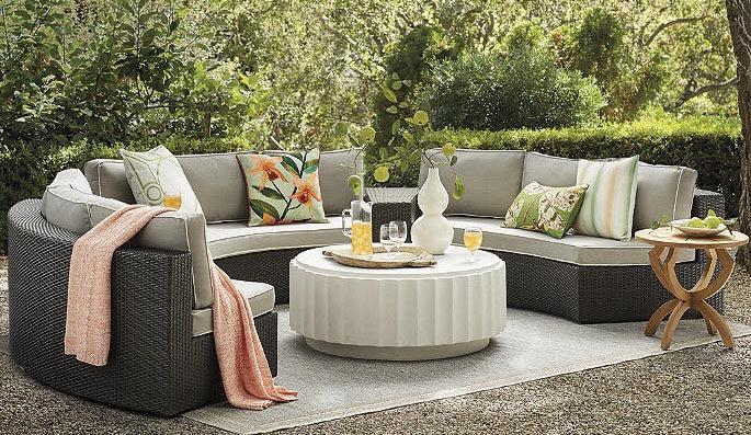 A cozy outdoor seating area featuring a curved wicker sectional sofa with beige cushions, colorful throw pillows, a white textured round coffee table with drinks and a decorative vase, a small wooden side table, and greenery in the background.