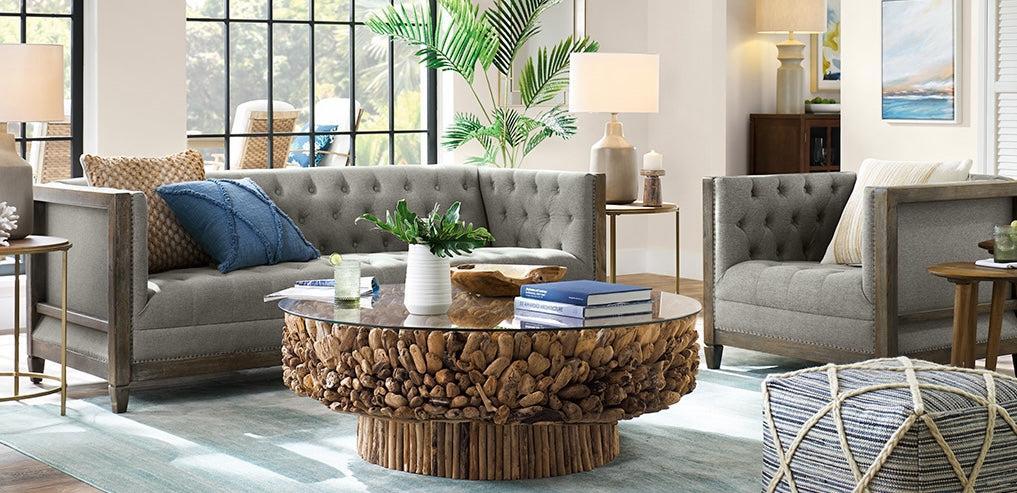 A modern, cozy living room with two gray tufted sofas, a unique wooden coffee table topped with books and plants, and various cushions. The room is bright with large windows, indoor plants, floor lamps, and a patterned pouf. A blue and white rug covers the floor.