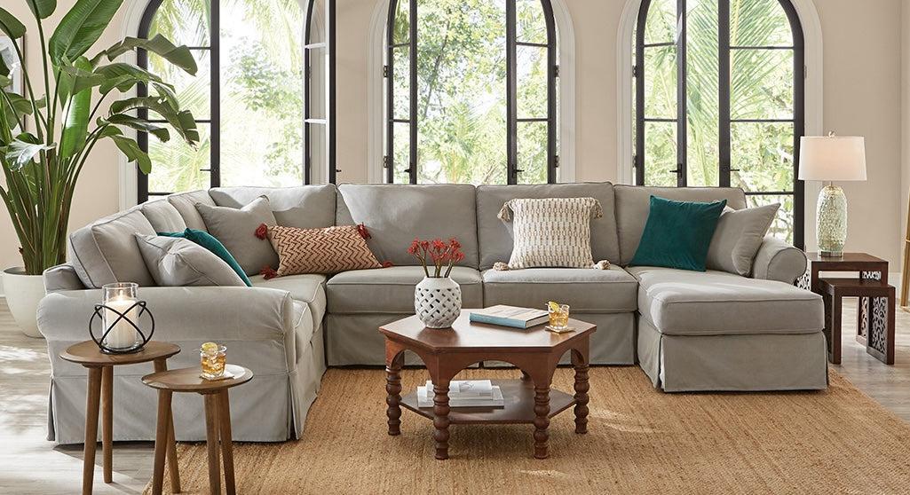 A spacious, elegantly decorated living room featuring a large gray sectional sofa with assorted colorful pillows. There are three wooden side tables, a central coffee table with décor items, and large arched windows allowing natural light to flood the space.