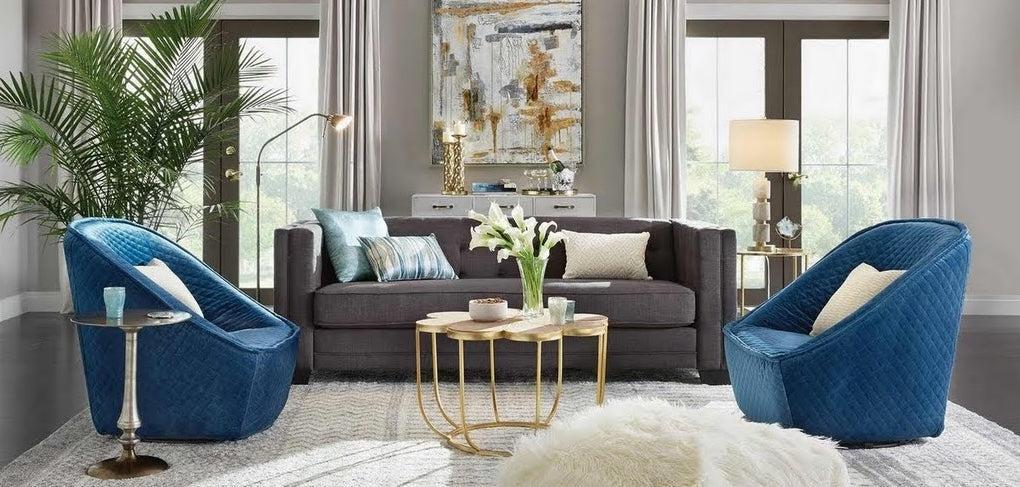 A modern living room featuring a gray sofa, two blue swivel chairs, and a gold coffee table with white flowers. The room is decorated with a large abstract painting, a tall floor lamp, gold accents, and a plush white rug. Light streams in from large windows with sheer curtains.