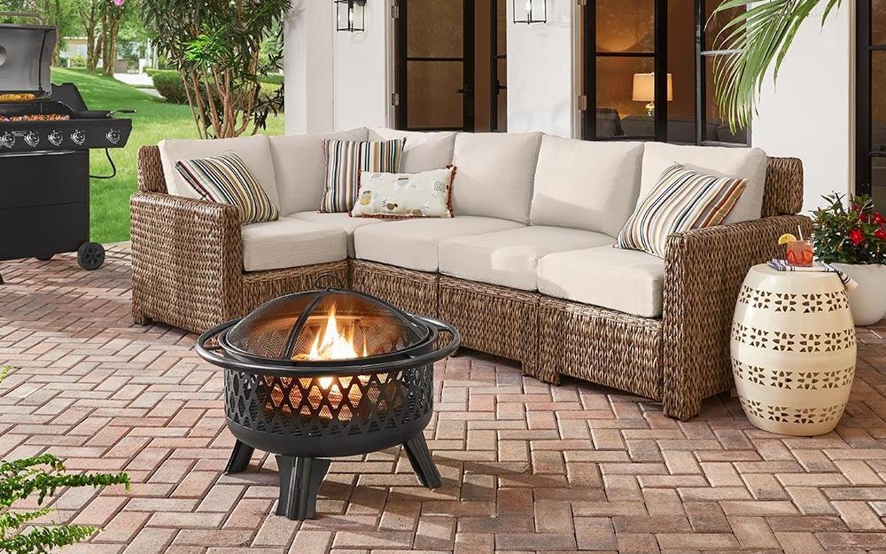 An outdoor patio features a wicker sectional sofa with beige cushions and striped accent pillows. A small fire pit burns in front of the seating area. There is a grill in the background and a decorative side table on the right. Greenery surrounds the patio.
