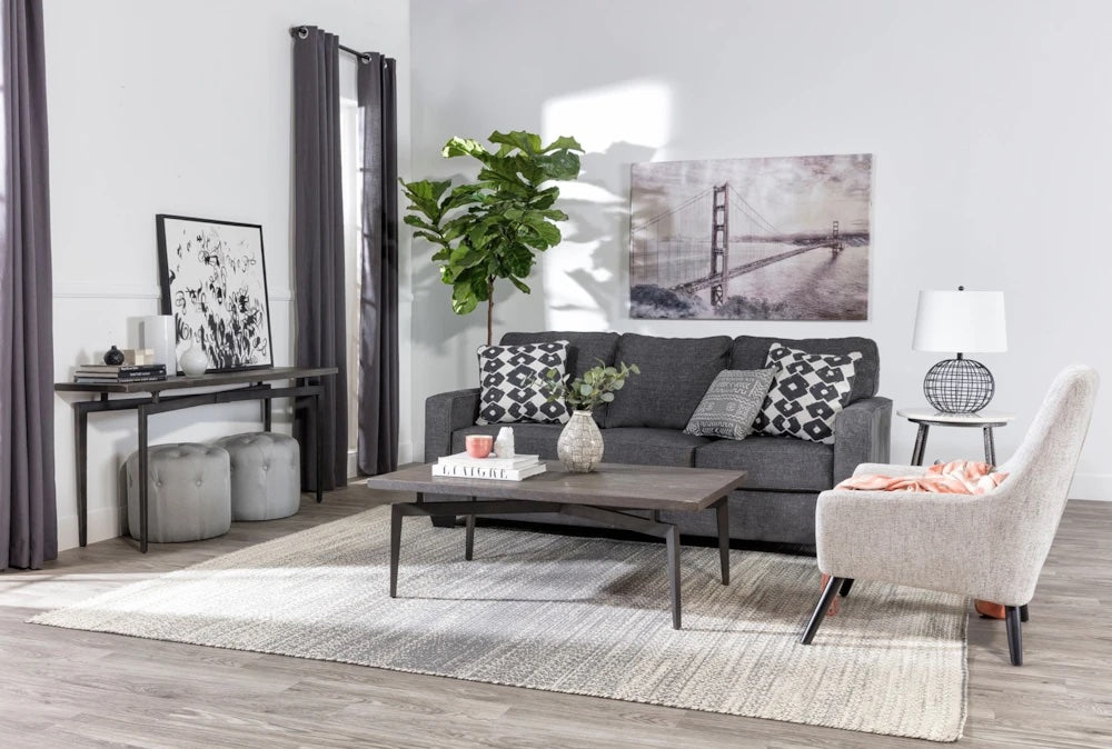 A modern living room with a gray sofa adorned with black and white patterned cushions, a light-colored armchair, and a coffee table on a textured rug. A tall green plant is in the corner, while wall art featuring a bridge hangs above the sofa.