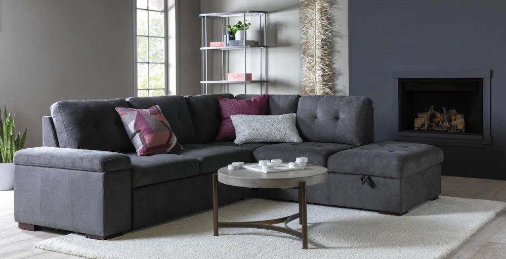 A modern living room featuring a dark gray sectional sofa with various cushions in shades of pink, purple, and gray. In front of the sofa is a round coffee table with white coordinating decor. The room includes a bookshelf, a fireplace, and a large window.