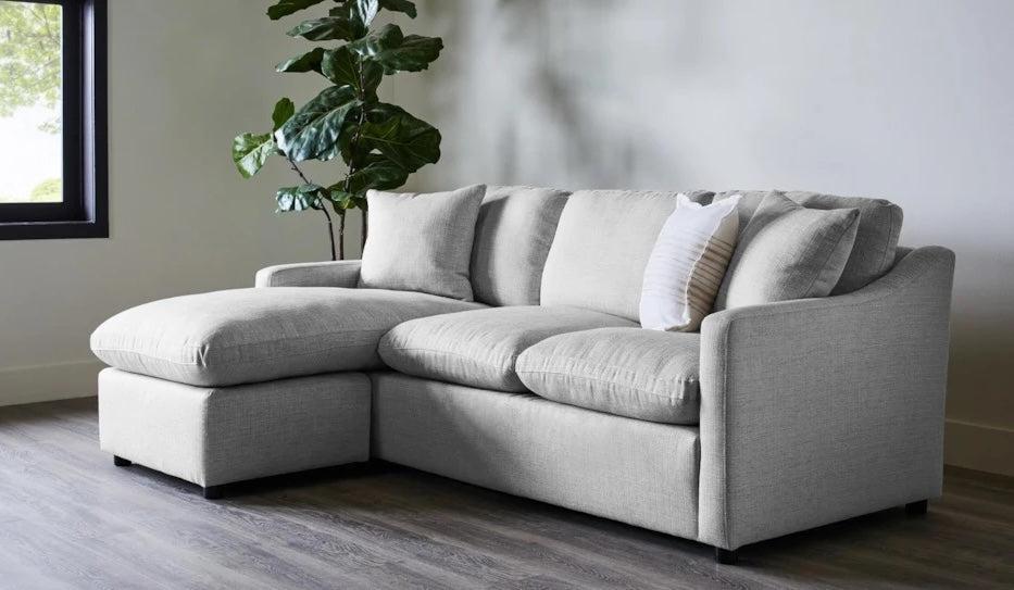 A modern living room features a light gray sectional sofa with a chaise lounge on the left side. A white and gray striped pillow is placed on the sofa. Behind the couch, a large green plant adds a touch of nature. The room has a minimalist design with wooden flooring.