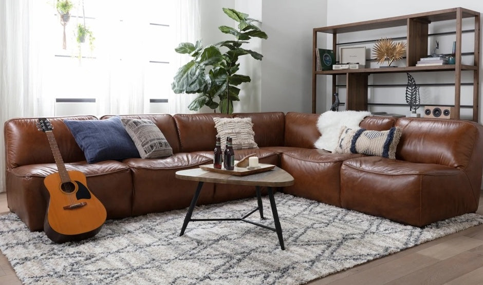 A cozy living room with a brown leather sectional sofa adorned with patterned and plain pillows. A guitar rests against the sofa. A small coffee table holds two bottles. A geometric rug covers the floor. A bookshelf with decorative items and a potted plant are in the background.