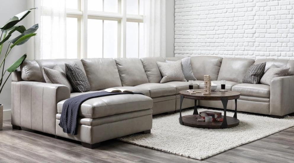 A modern living room with a large, light grey sectional sofa adorned with various throw pillows and a dark blue blanket. A round wooden coffee table with decor items sits on a light-colored rug. Behind the sofa, there's a white brick wall and large windows.