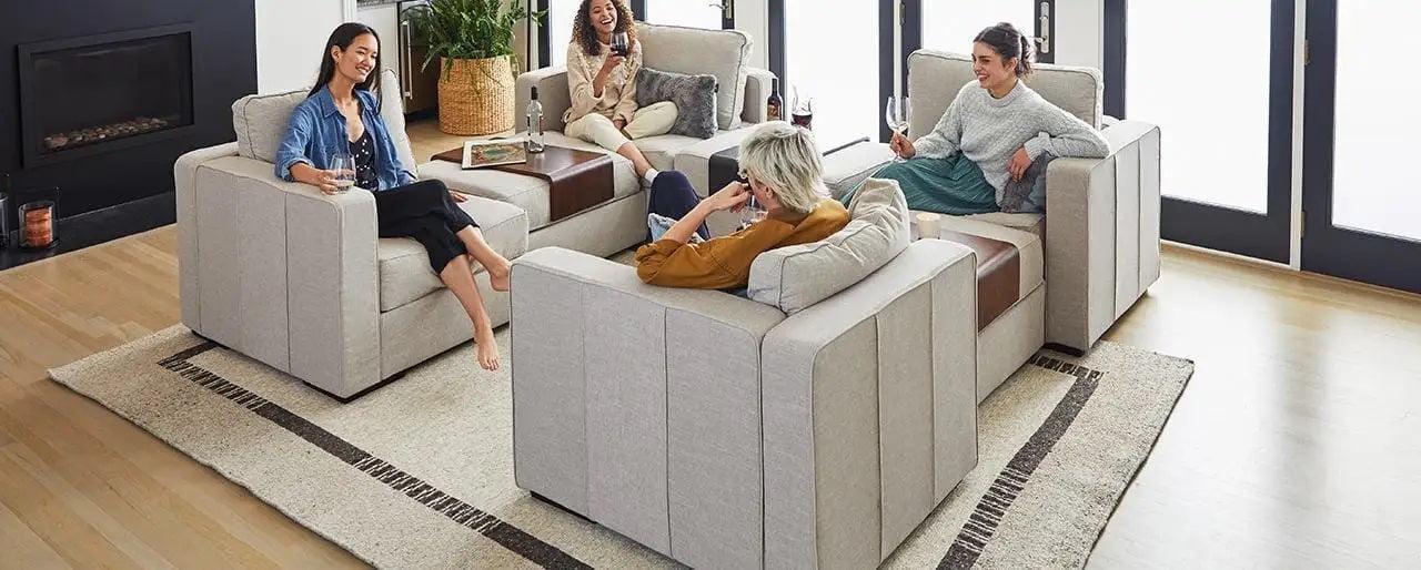 Four people sit on beige couches placed around a wooden coffee table in a modern living room with hardwood floors. A rug lies beneath the furniture, and natural light enters through large windows. They appear to be engaging in a pleasant conversation.