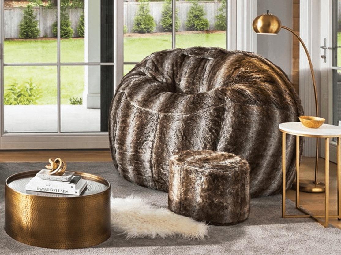 A cozy living room setup featuring a large faux fur bean bag chair, a matching faux fur ottoman, a round metallic coffee table with books and a decorative knot, a white furry rug, and a gold floor lamp. Large windows in the background allow natural light in.