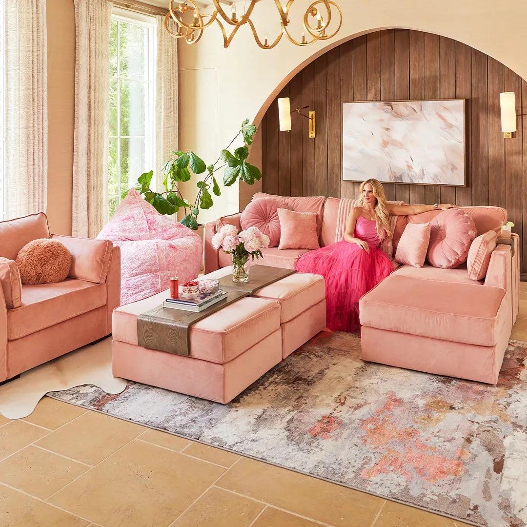 A bright living room features a sectional sofa, armchairs, a sizable ottoman, and a bean bag chair, all in a soft pink hue. A woman in a flowing pink dress sits on the couch. The room is adorned with plants, a wall painting, and a decorative chandelier.