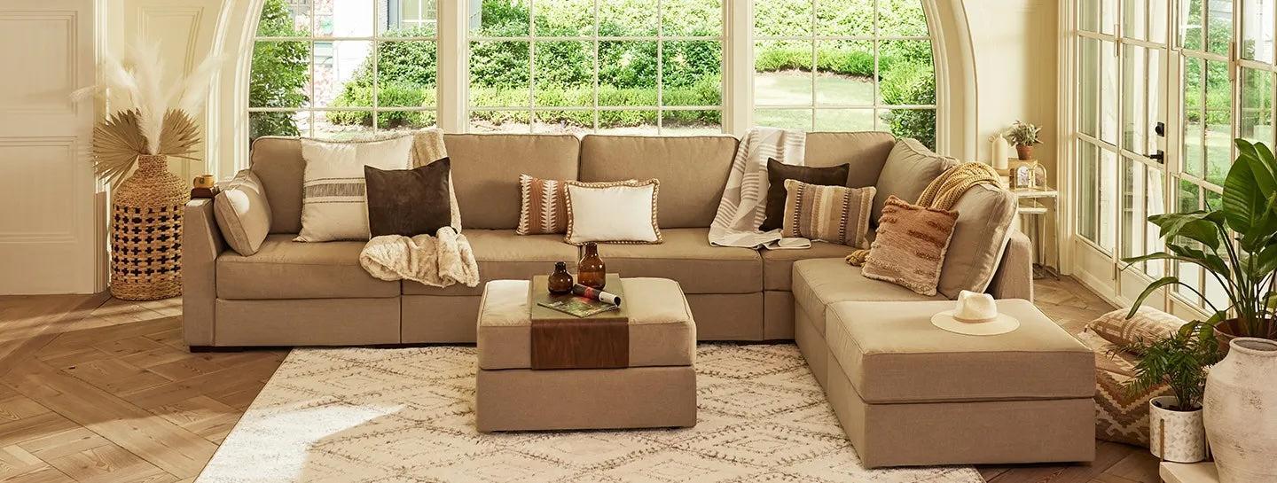 A cozy living room with a large beige sectional sofa decorated with various pillows and throws. An ottoman serves as a coffee table with decorative items on it. The space is well-lit with natural light from the large windows, and plants and decor accents embellish the scene.