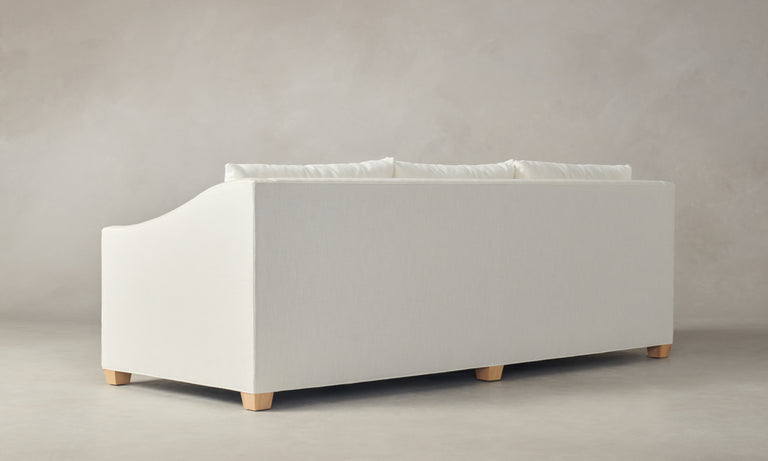 The Sullivan 60" Sofa in Performance Linen Weave, featuring a minimalist and modern design with a soft fabric cover in white and wooden legs, is set against a neutral gray background. The sofa's back is visible, highlighting its simple and clean aesthetic.