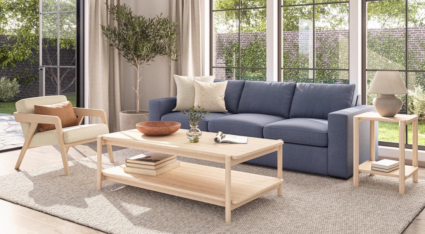 A bright living room with large windows overlooking a green garden. The room features a blue sofa with decorative pillows, a wooden coffee table with a vase and books, a beige armchair, a side table with a lamp, and a potted plant in the corner.