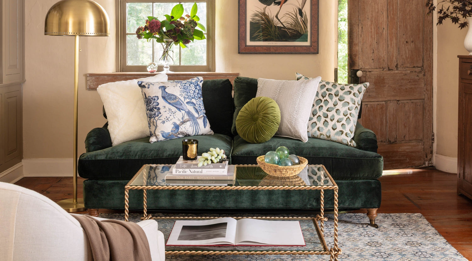 A cozy living room features a dark green velvet sofa adorned with various patterned and textured throw pillows. A gold floor lamp stands beside it. A glass coffee table with books, decor, and a bowl of green spheres sits on a patterned rug. Soft natural light fills the room.