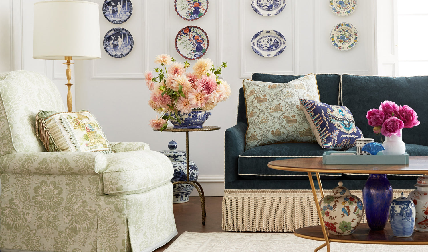 A cozy living room features a cushioned armchair with a green floral pattern, a dark green sofa with decorative pillows, and a wooden coffee table with a vase of pink flowers. Blue and white decorative plates adorn the wall, and a side table holds pastel flowers.