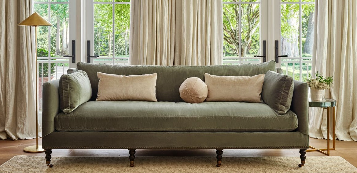 A cozy living room featuring a green upholstered sofa with two rectangular cream pillows and one round pink pillow. The sofa is framed by large windows with beige curtains. A gold floor lamp stands to the left and a small plant on a stand is to the right.