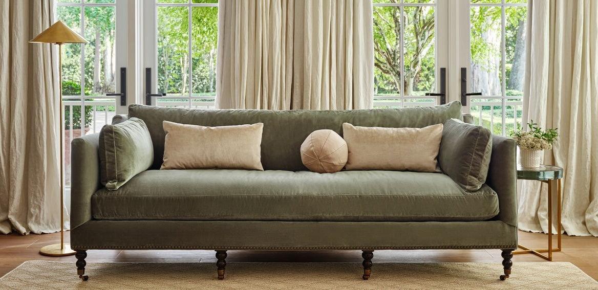 A cozy living room featuring a green upholstered sofa with two rectangular cream pillows and one round pink pillow. The sofa is framed by large windows with beige curtains. A gold floor lamp stands to the left and a small plant on a stand is to the right.