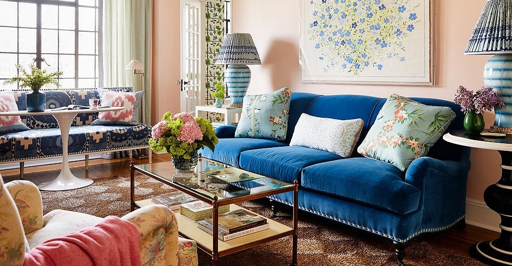 A vibrant living room features a plush blue sofa adorned with floral pillows, a glass coffee table with books and flowers, patterned armchairs, and a floral print on the wall. Two striped lamps sit on side tables, and a large window lets in natural light.